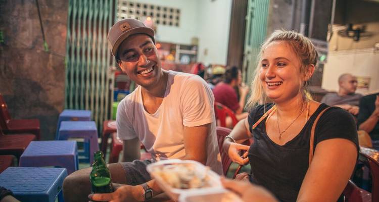 Smiling friends enjoy beer and street food at a casual night eatery with plastic stools.