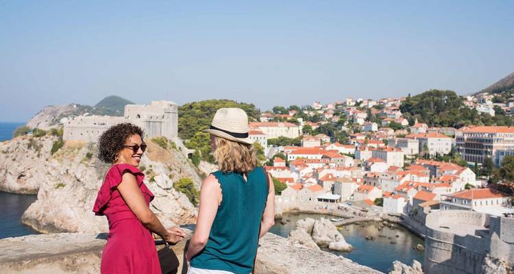 Dos mujeres observando un pintoresco pueblo costero con fortificaciones históricas de piedra.