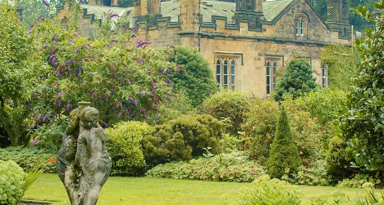 Jardin de domaine historique avec une végétation dense, des plantes à fleurs et une façade de maison ancienne en pierre.