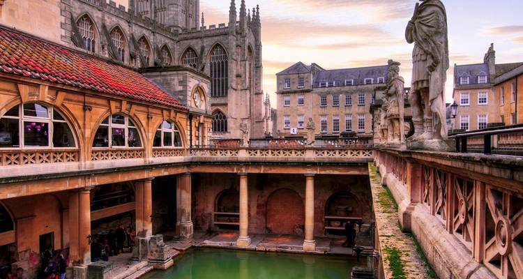 Complexe historique de thermes romains avec statues et bassin thermal illuminé par un ciel de coucher de soleil aux couleurs pastel.