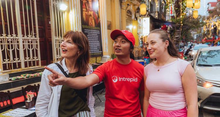 Guía local con camiseta roja señala lugares de interés a dos viajeros en una calle animada de Hanói al atardecer