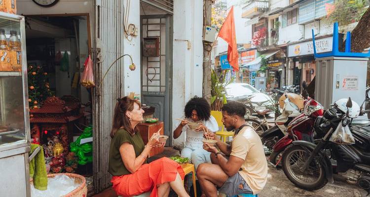 Los viajeros se sientan en banquitos bajos disfrutando de fideos en un pequeño puesto callejero de Hanoi rodeado de motos estacionadas