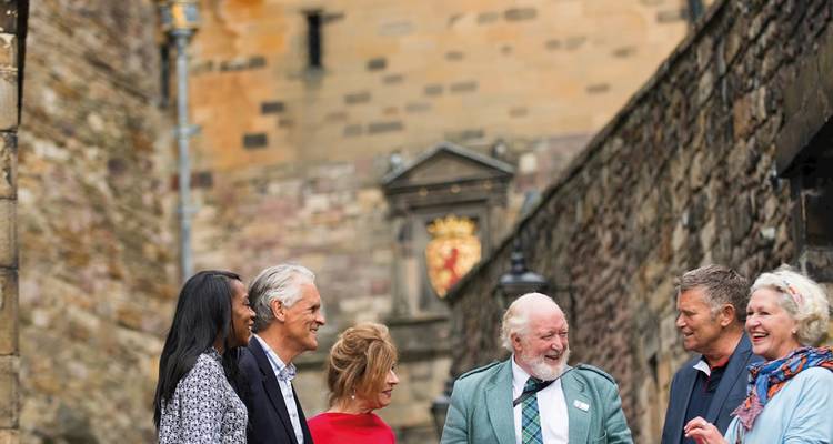 Un grupo mixto conversa afuera de una muralla de castillo de piedra adornada con un escudo real en Escocia.