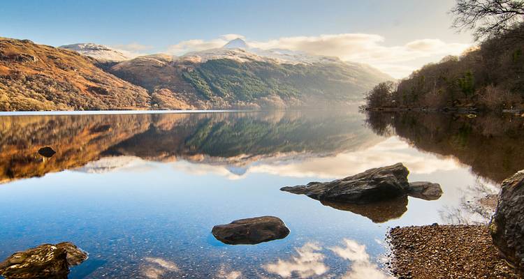 Lago inmóvil como cristal que refleja las colinas boscosas circundantes y un pico nevado al amanecer