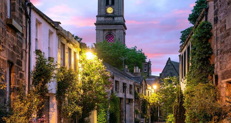 Charmante ruelle étroite en pierre bordée de maisons couvertes de lierre menant vers un clocher sous un ciel de coucher de soleil coloré.