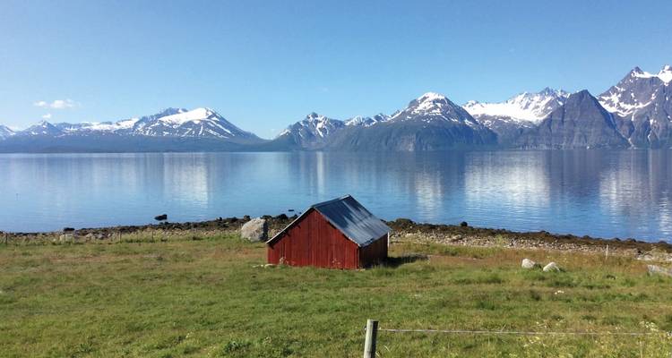 Idyllic landscape with a red cabin by a lake and snowy mountains.
