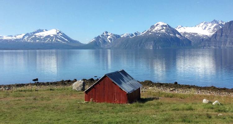 Casa de botes roja solitaria en orilla herbosa con aguas tranquilas del fiordo y picos nevados al fondo