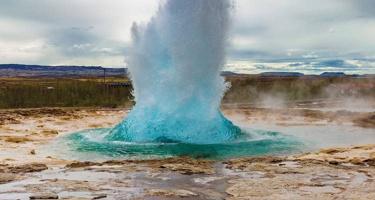 Puissante éruption de geyser bleu jaillissant vers le ciel au milieu d'un paysage géothermique islandais.