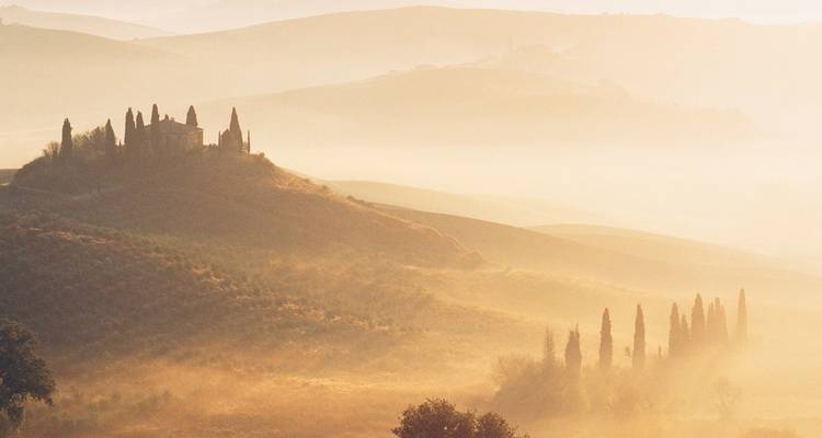 La brume matinale dorée s'élève sur les collines toscanes en étages, avec une ferme perchée sur une colline et des rangées de cyprès.