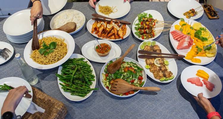 Bovenaanzicht van een overvloedige verscheidenheid aan Aziatische gerechten en gegrilde spiesjes op een tafel terwijl verschillende gasten erin reiken met serveerlepels
