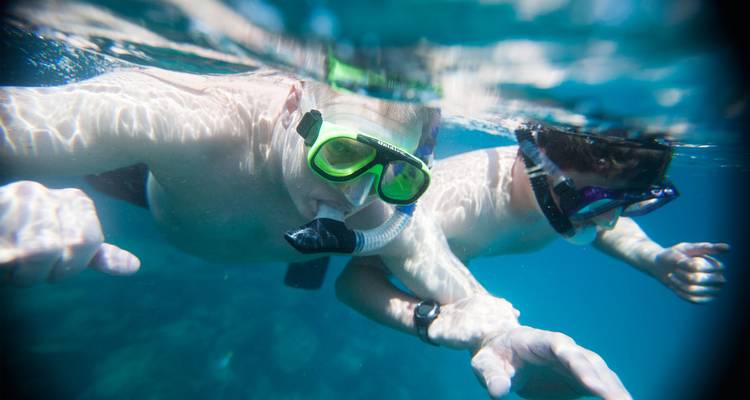 Twee snorkelers met maskers en snorkels zwemmen boven helder blauw water, zonlicht dat op hun huid golft.