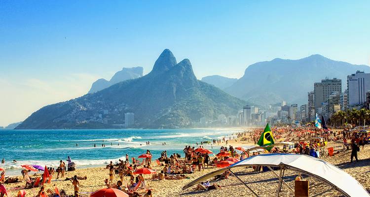 Playa de Ipanema abarrotada con sombrillas, banderas brasileñas y montañas dramáticas elevándose detrás del horizonte de la ciudad.