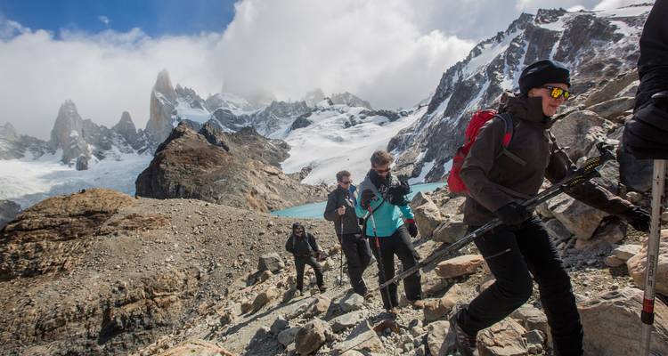 Los escaladores atraviesan una cresta rocosa bajo picos nevados irregulares y un lago glacial color turquesa en la Patagonia.