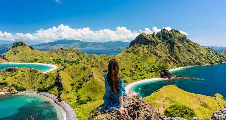 Een reiziger in een blauwe jurk zit op een klif en bewondert de levendige groene heuvels en blauwe lagunes van Komodo National Park.
