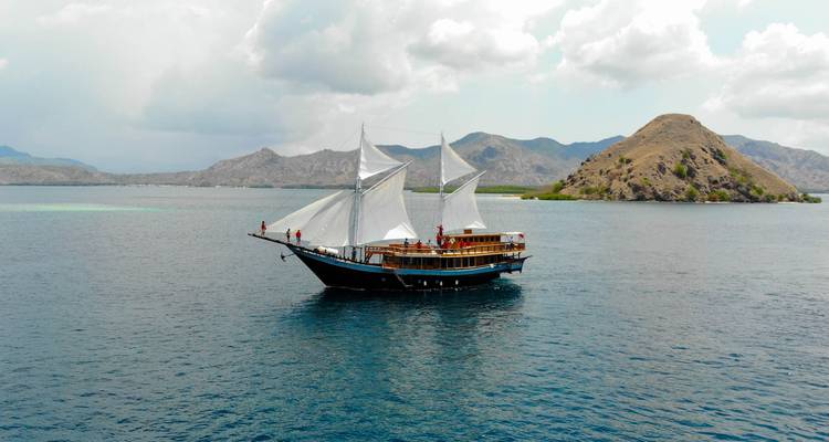 Traditional two-masted wooden phinisi sailing on calm blue waters with rugged islands in the distance