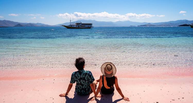 Young couple sitting on a pink sand beach gazing at a anchored boat on clear blue sea