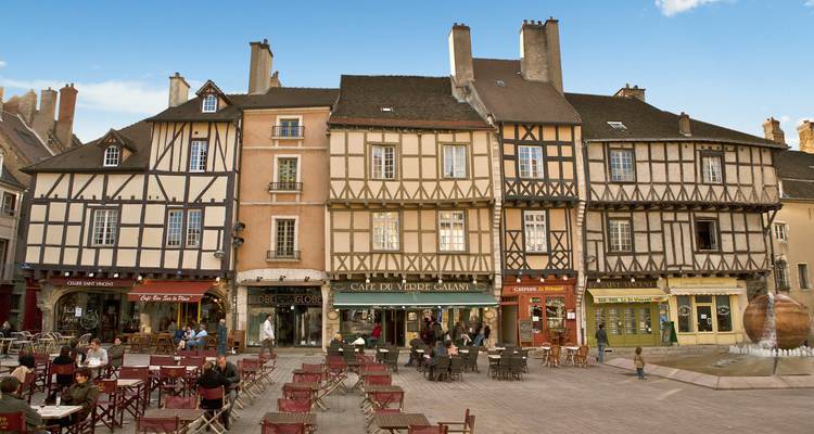 Edificios tradicionales franceses que rodean una animada plaza con asientos al aire libre.
