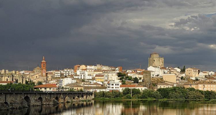 Historic town with stone bridge and fortress tower under a dramatic dark sky reflected in a river