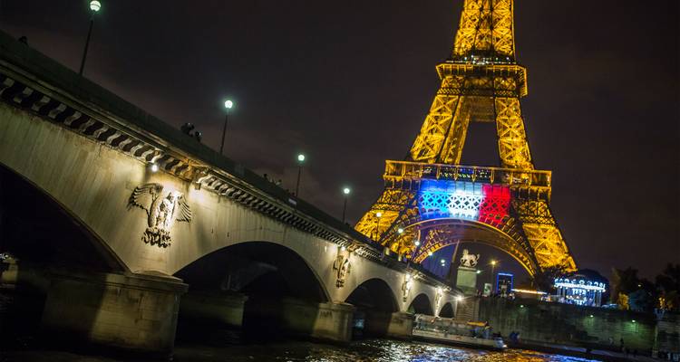 Night-time view of the Eiffel Tower lit in gold with French flag colors under a bridge.