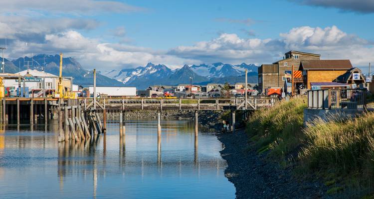 Harbor scene with wooden pier, buildings and snow-dusted mountains under blue sky in Homer