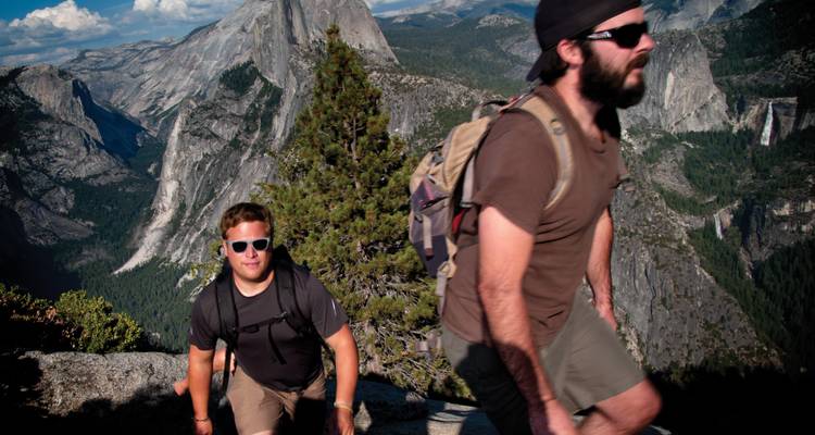 Zwei Wanderer mit Rucksäcken, die einen felsigen Weg im Yosemite erklimmen, mit dramatischen Granitgipfeln im Hintergrund.