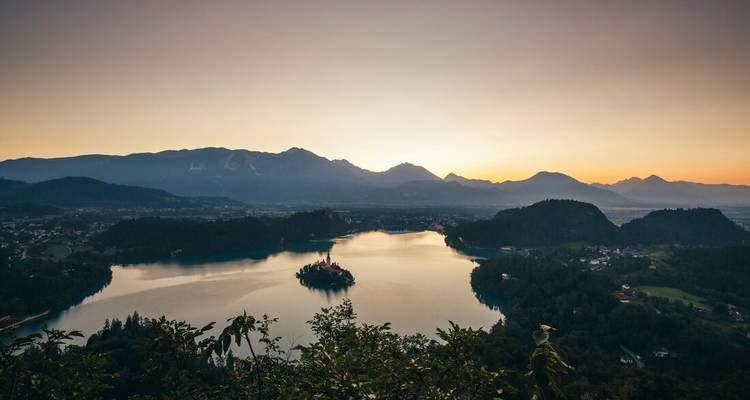 Sunrise casts warm hues over Lake Bled and its island church framed by forested hills and distant Alps.
