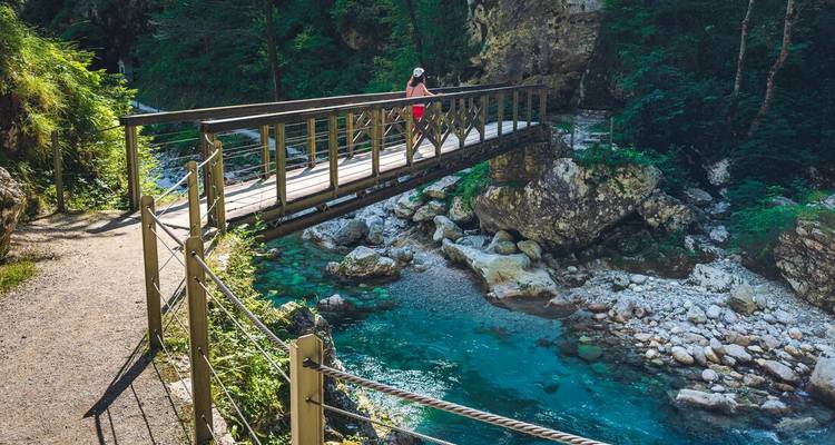 A hiker crosses a wooden bridge over crystal-clear turquoise water in a lush forest gorge.
