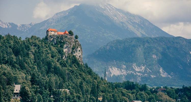 Bled Castle perched on a rocky cliff stands before misty Alpine peaks under a partly cloudy sky.