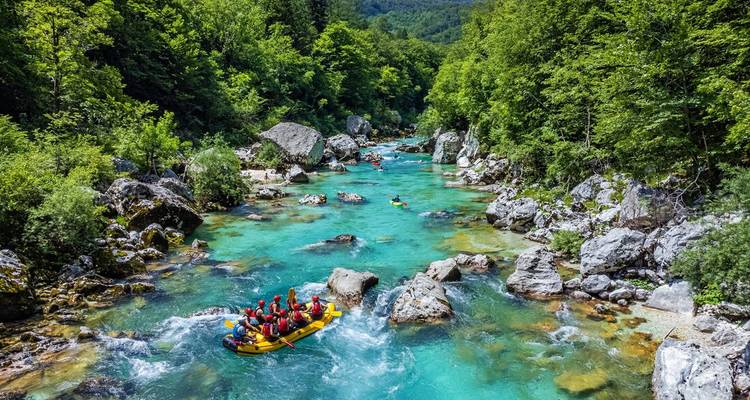 An inflatable raft packed with paddlers navigates the vivid turquoise rapids of the Soča River through lush forest.