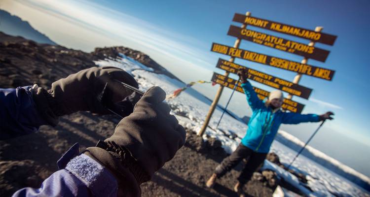 Ein triumphierender Bergsteiger feiert am Schild des Uhuru Peak, während ein Begleiter ein Foto macht.
