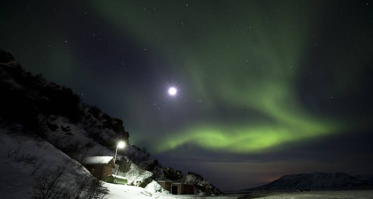 Aurora Boreal sobre un paisaje nevado con una cabaña iluminada por la luna.