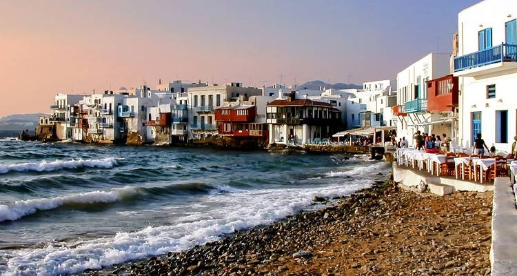 Paseo marítimo de la Pequeña Venecia de Mykonos con balcones coloridos al atardecer, olas rompiendo en la playa de guijarros.