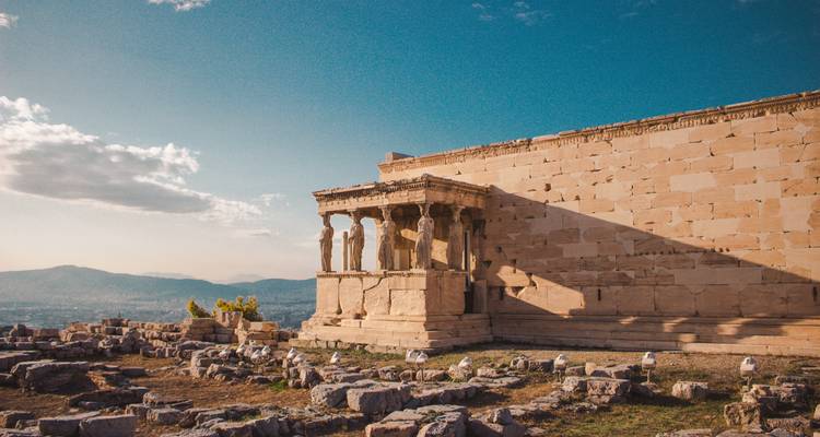 Sun-kissed Erechtheion with its famous Caryatid statues overlooking rocky ruins and distant hazy mountains.