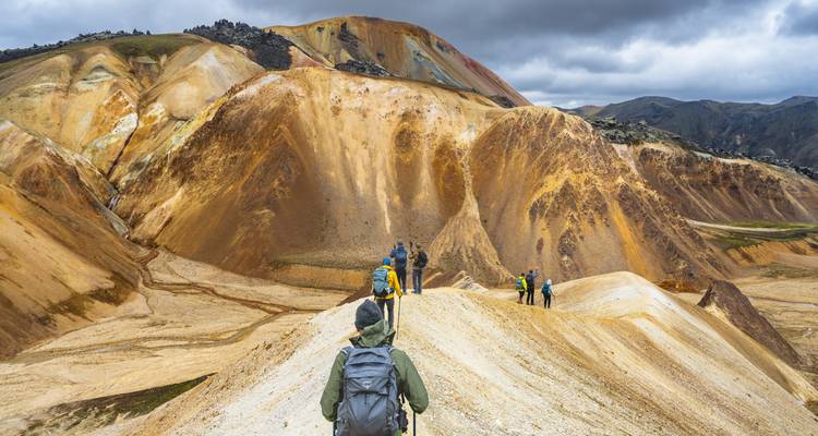 Fila de excursionistas con mochilas atraviesa una cresta estrecha color crema entre dramáticas colinas volcánicas ocres.