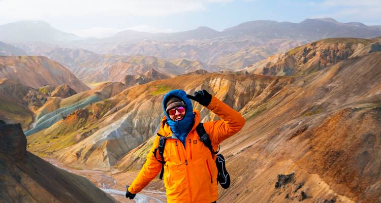 Excursionista alegre con chaqueta naranja brillante saluda desde una cresta volcánica azotada por el viento con colinas coloridas detrás.