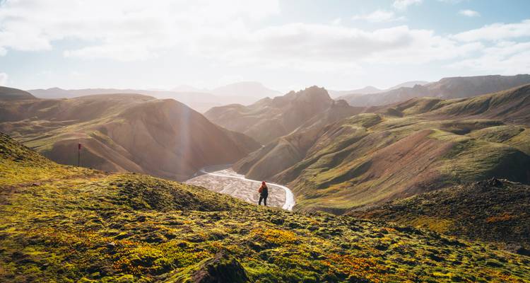 Un excursionista solitario se encuentra en un valle iluminado por el sol con crestas dramáticas y un río serpenteante que brilla bajo los rayos de la tarde.