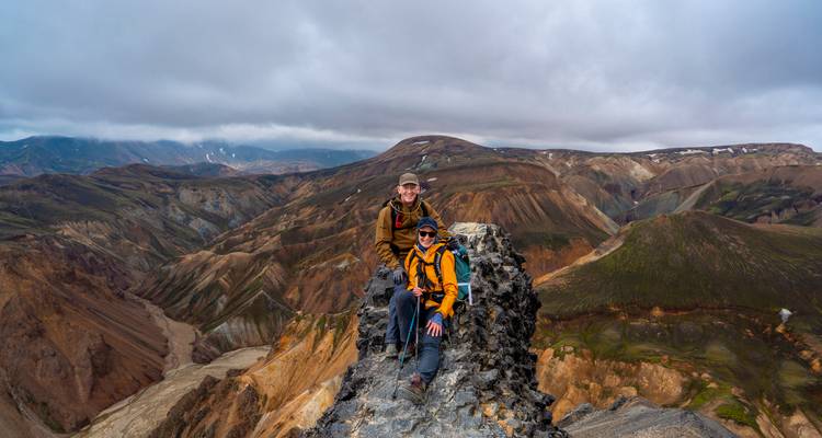 Two hikers perch on a rocky pinnacle overlooking vast colourful valleys.