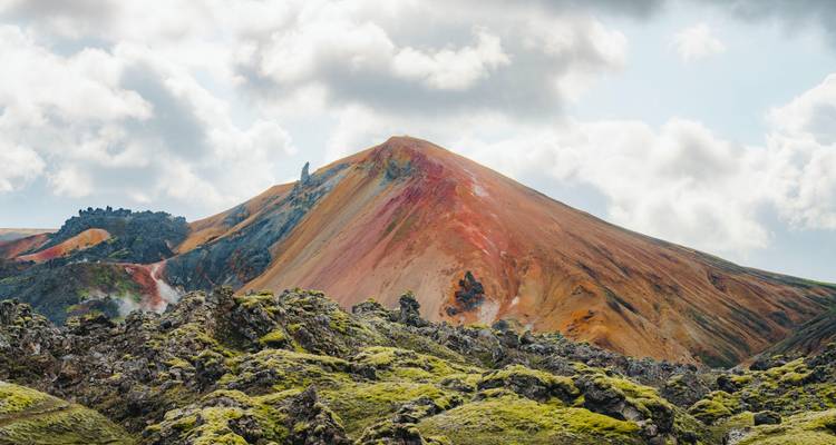 Rust-red rhyolite peak framed by mossy lava rocks under a bright sky.