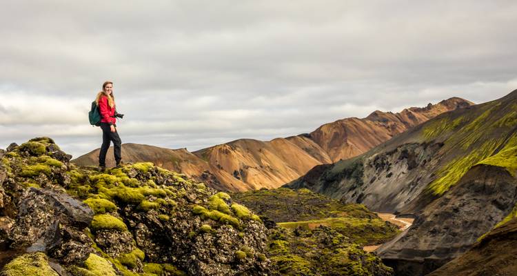Solo traveller stands atop moss-covered volcanic rock with dramatic orange mountains behind.
