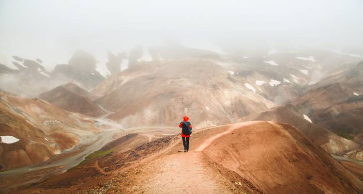 Hiker walks a fog-shrouded volcanic ridge amid reddish earth and snow patches.