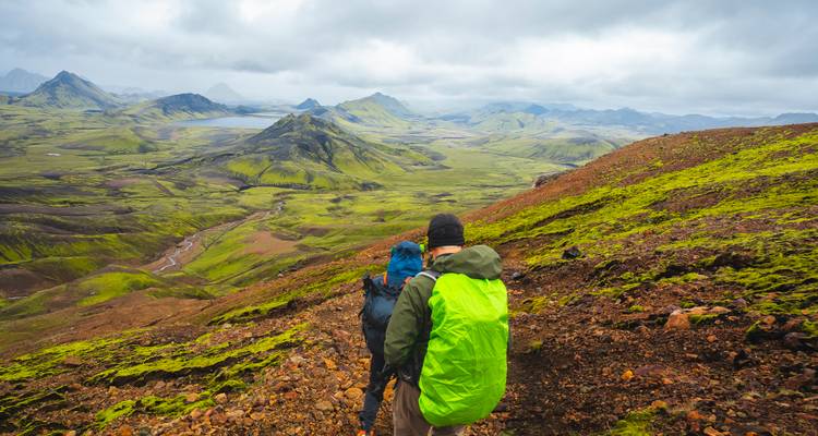 Hikers descend a moss-covered slope overlooking lakes and volcanic valleys.