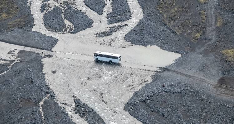 White bus fords a braided glacial river across a rocky highland plain viewed from above.