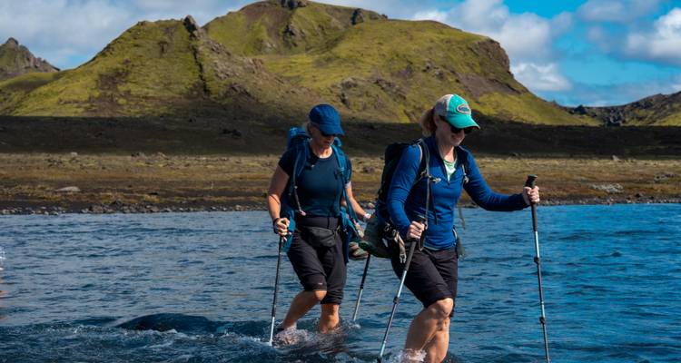 Deux randonneurs traversent une rivière à hauteur des genoux avec des bâtons, une crête volcanique verte sous un ciel bleu en arrière-plan.