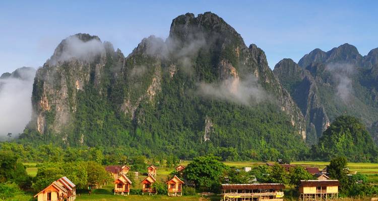Des imposantes montagnes karstiques couvertes de brume s'élevant derrière un petit village rural à Vang Vieng.