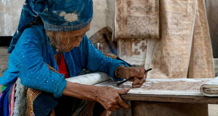 Un artesano anciano dibuja cuidadosamente patrones en tela mientras está sentado junto a textiles tradicionales.
