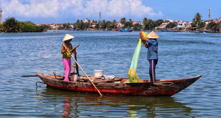 Zwei vietnamesische Fischer in Kegelhueten werfen ein Netz von einem Holzboot auf einem ruhigen Fluss aus.