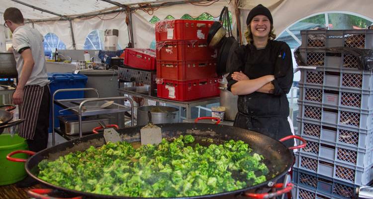 Chef sonriendo junto a una gran sartén de verduras en una cocina al aire libre.