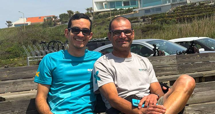 Dos guías sonrientes se relajan en un banco de madera con bicicletas y villas junto al mar detrás.