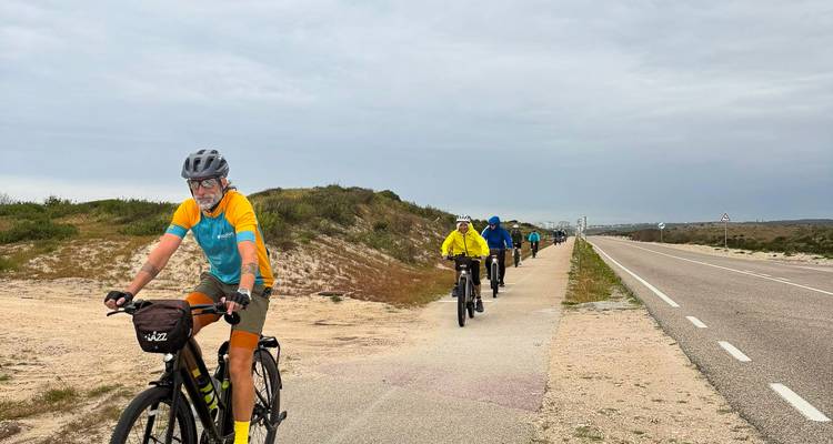 Los ciclistas avanzan en fila india por un carril bici costero azotado por el viento bajo un cielo nublado.
