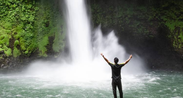 Männlicher Reisender mit erhobenen Armen vor einem beeindruckenden Dschungelwasserfall.